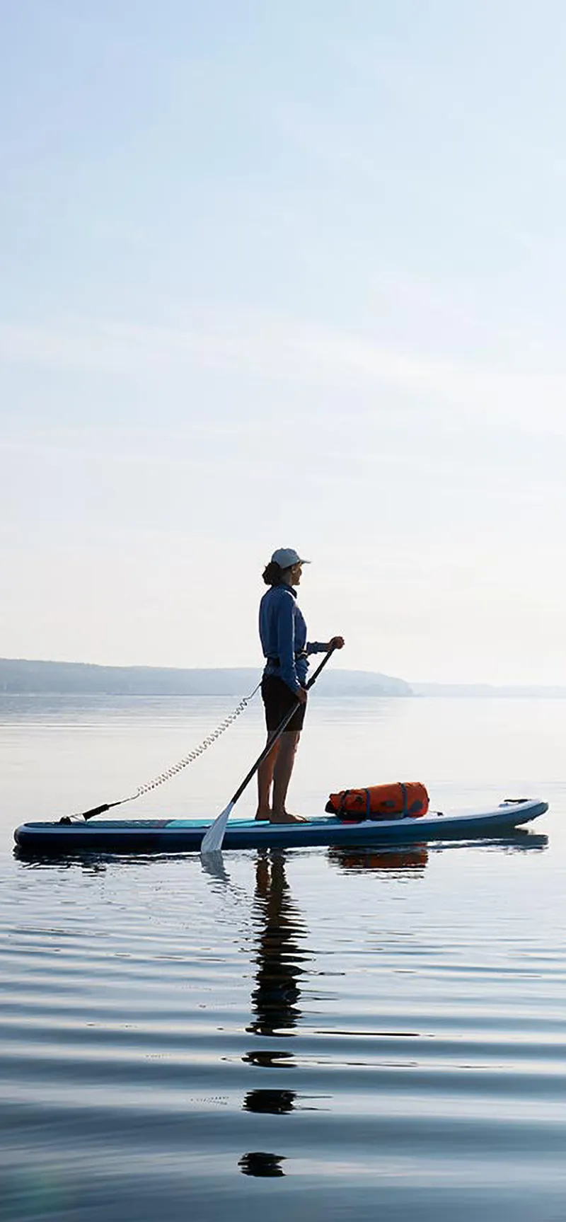 Frau steht auf einem stand up paddle board und paddelt auf einem See, die Sonne scheint